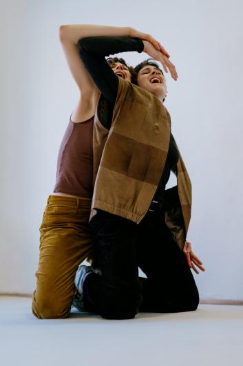 Two people lean closely into each other, their bodies intertwined in a flowing movement; one wears a brown vest, both appear to laugh or call out.