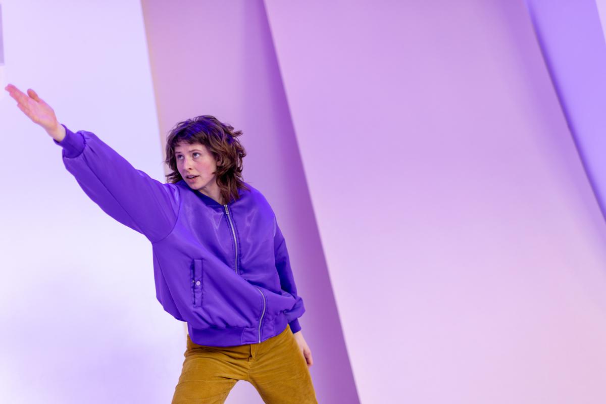 A person in a purple jacket extends an arm forward, body slightly tilted, against a geometrically structured wall under purple lighting.