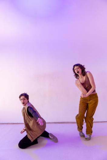 Two people move separately in space, one kneeling on the floor, the other standing; both engaged in intense, expressive movement under purple light.