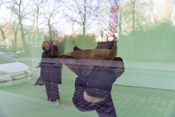 Durch eine große Glasfläche fotografiert: Im Vordergrund dreht sich eine Person in einer braunen Weste und dunkler Kleidung, ein Arm ist angehoben und die Weste fliegt seitlich auf. In der Spiegelung links stehen zwei weitere Personen unscharf im Raum. Draußen hinter der Glasfront sind ein geparktes Auto, kahle Bäume und der Gehsteig zu sehen.