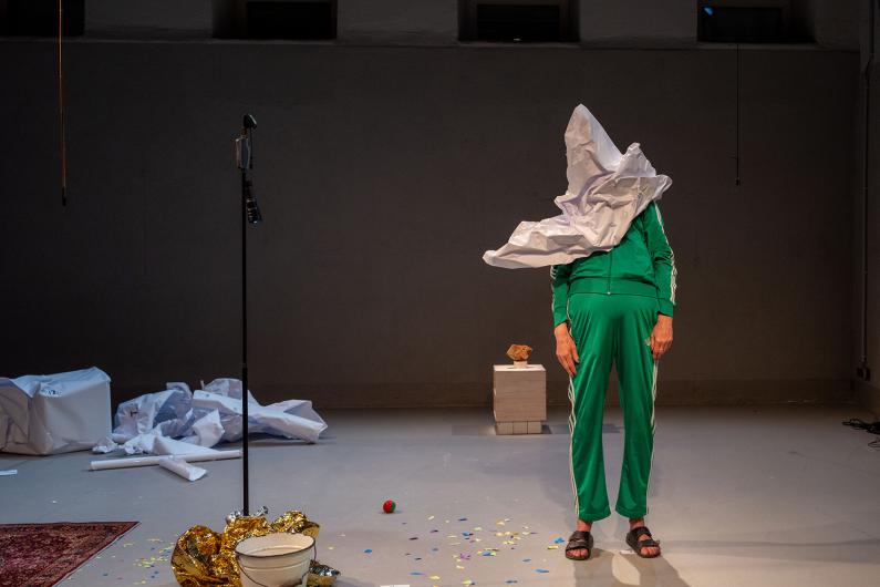 A person in a green tracksuit stands facing forwards on stage, their head completely hidden by a large crumpled white paper shape. To the left are a microphone stand and a white bowl, with confetti and props scattered on the floor. The scene feels still, absurd and slightly unsettling.