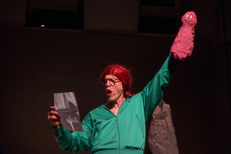 Close view of a person on a dark stage wearing a green tracksuit and a red wig. In one hand they hold a sheet of paper, in the other a roughly shaped pink figure with a drawn face. Their mouth is wide open, as if speaking or calling out