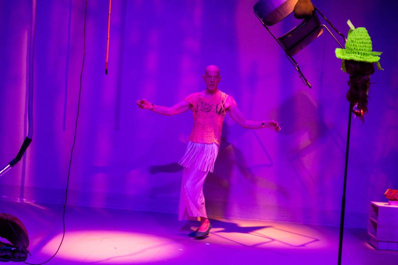 A person stands alone in violet-blue stage light, moving through the space with arms extended. They wear a delicate pale top and a long white fringed skirt. Cables and props hang down from above, giving the scene the feel of an installation.