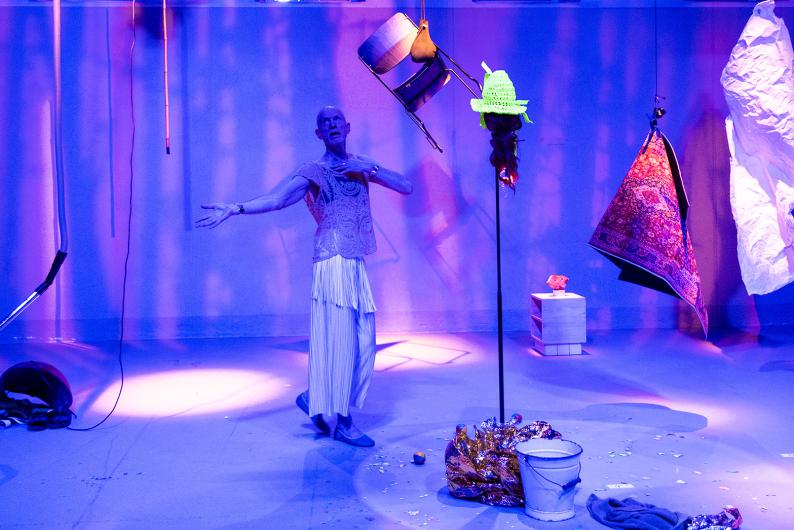 A person in a pale stage costume stands in blue-violet light with one hand on their chest and the other arm opened to the side. Beside them hang a green crocheted headpiece with hair and a rug suspended at an angle. On the floor are a white bucket, shimmering fabric and small balls.