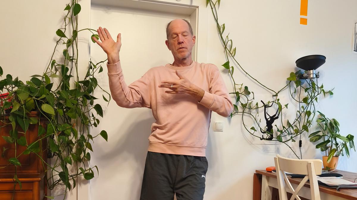 A person stands in a bright living room in front of a white door, eyes closed, with their arms raised in a calm, gestural pose. Their left hand is open and lifted upwards, while the right hand hovers in front of the chest. Green houseplants trail across the wall around the door. On the right, there is a small table with a chair, papers and more plants, along with a black wall lamp. The person is wearing a pale pink sweatshirt and dark jogging bottoms. The scene feels quiet, focused and intimate.