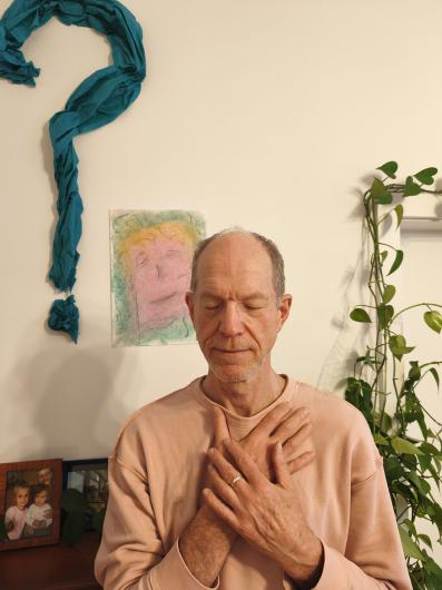 An older adult with light skin sits in front of a living-room wall with plants and children’s photos. Above the person hangs a large turquoise fabric question mark and a colourful portrait drawing. The person wears a pink sweatshirt, eyes closed, with both hands crossed over the chest, as if breathing calmly or meditating.