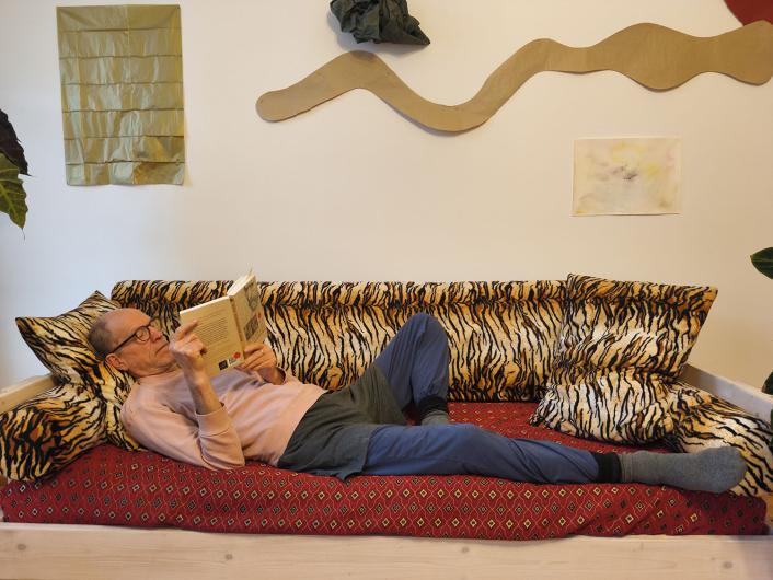 Eine älter werdende Person mit heller Haut liegt quer auf einem breiten Sofa mit Tigerfell-Muster und roter gemusterter Unterlage. Die Person trägt ein rosa Sweatshirt, blaue Hosen und graue Socken und hält ein aufgeschlagenes Buch vor sich, konzentriert lesend. Hinter dem Sofa hängt abstrakte Wanddekoration in Gold- und Beigetönen.