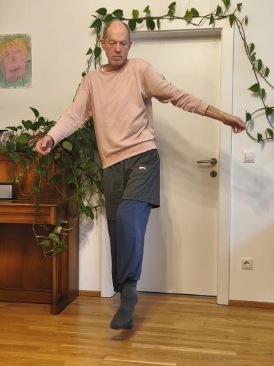 Eine älter werdende Person mit heller Haut steht barfuß im Wohnzimmer auf einem Holzfußboden vor einer weißen Tür, umrahmt von vielen grünen Rankpflanzen. Die Person trägt ein rosa Sweatshirt, weite dunkelblaue Hosen mit grauen Shorts darüber und graue Socken. Ein Arm ist zur Seite ausgestreckt, ein Bein angehoben – die Haltung wirkt wie eine tänzerische Balancebewegung.