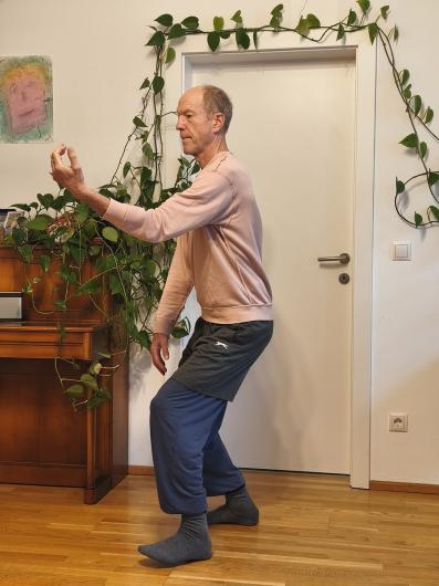 Die gleiche älter werdende Person mit heller Haut steht im Wohnzimmer vor der Tür, umgeben von Topfpflanzen auf einem Klavier. Die Person trägt ein rosa Sweatshirt, graue Shorts über weiten blauen Hosen und graue Socken. In leicht gebeugter Haltung hält sie eine Hand vor dem Körper, als würde etwas betrachtet oder gehalten, die andere Hand hängt entspannt nach unten.