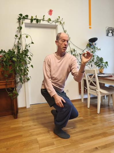 An older adult with light skin kneels on one leg on the wooden floor in the living room. The person is dressed in a pink sweatshirt, grey shorts over loose blue trousers and grey socks. In front of a white-framed door with green climbing plants, the person shapes a small gesture with one hand, as if carefully holding or examining something tiny.