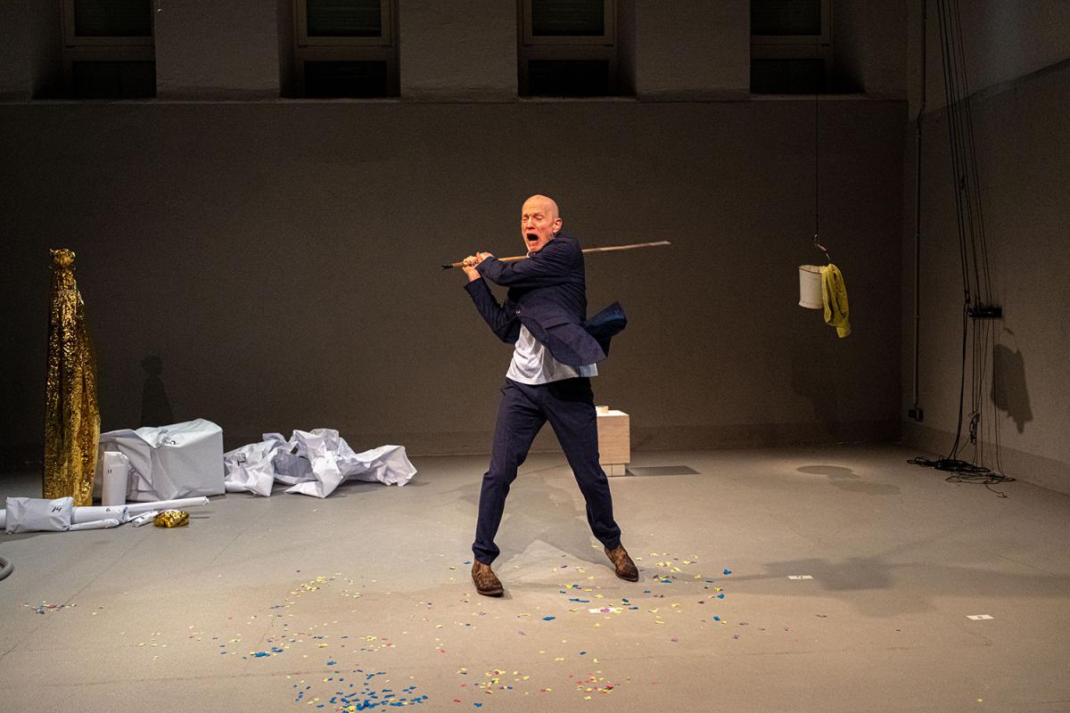 A person in a blue suit stands wide-legged on stage, swinging a long pole across the space. Their mouth is open and the movement feels forceful and dramatic. Confetti, crumpled white paper and a gold covering are scattered across the floor around them.