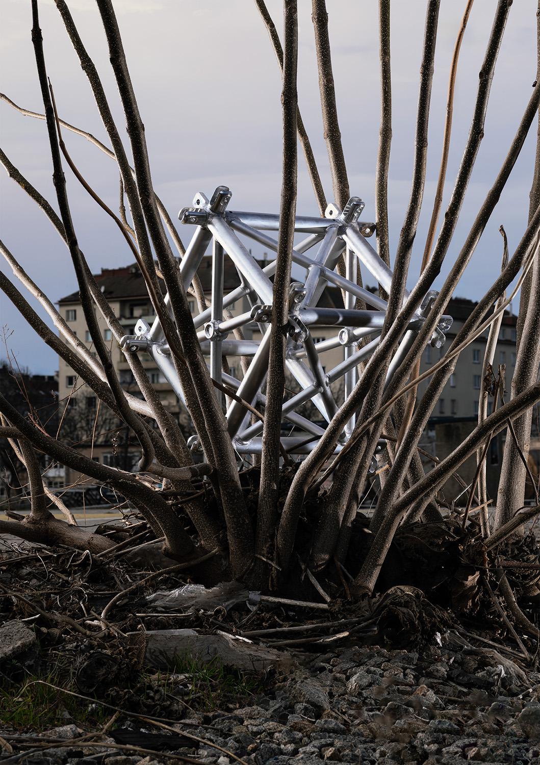 A silver geometric metal structure stands among dense bare branches. Building facades are visible in the background.