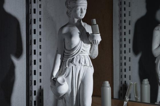 Eine beleuchtete weiße Statue in einem backstage-ähnlichen Raum steht vor einem Spiegel. Die Statue hält zwei moderne Flaschen in den Händen. Rundherum liegen Kämme, Bürsten und weitere Haar-Styling-Objekte. Links ist ein großer Schatten der Statue zu sehen. 