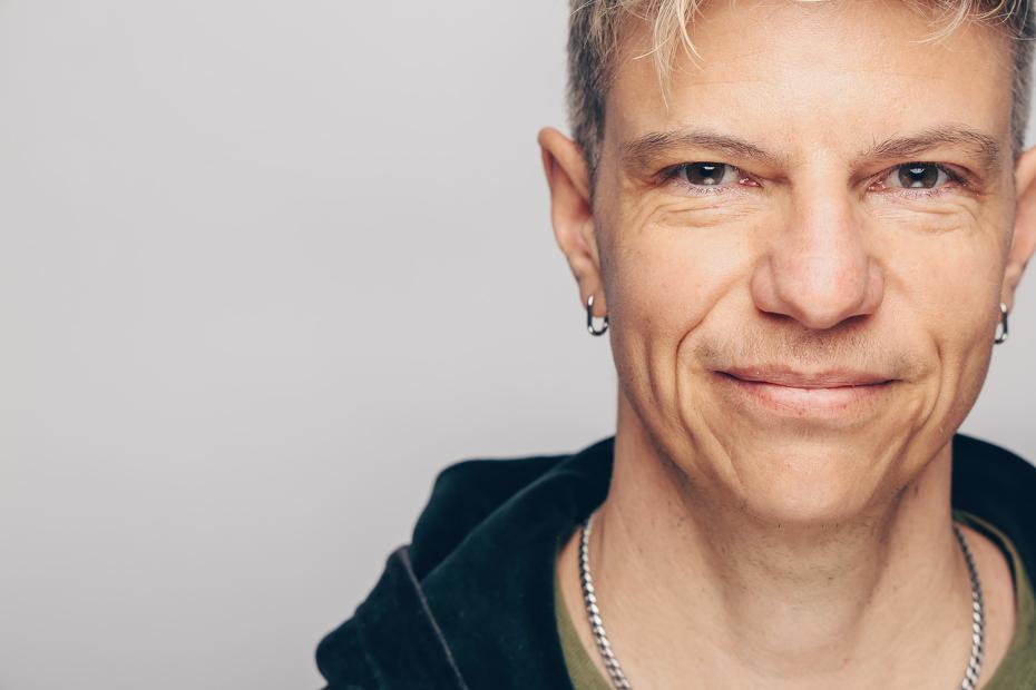 Nahaufnahme einer Person mit kurzem, blondem Haar, die leicht lächelt. Die Person trägt eine dunkle Jacke, ein grünes Oberteil und eine silberne Kette. Der Hintergrund ist neutral hellgrau.