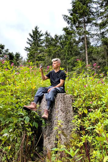 Eine Person mit kurzem Haar sitzt auf einem großen Baumstumpf inmitten von rosa blühenden Pflanzen und hält ein Smartphone nach oben, als würde sie fotografieren.
