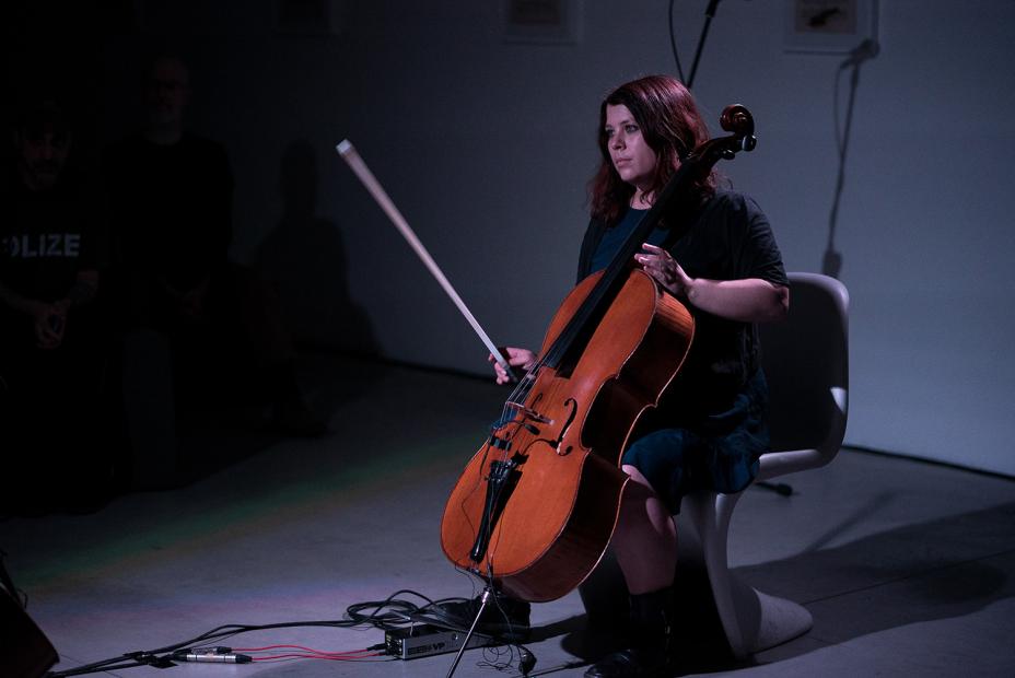 A person sits on a chair in a dimly lit room, playing a cello. The instrument is connected with cables, and several audience members sit in the background.