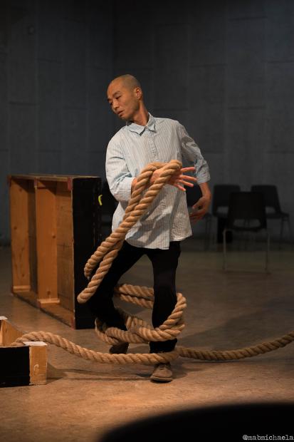 A person stands in a gray-walled room, dancing with a thick rope wrapped around their legs and arms. The person wears a light blue striped shirt and black pants.