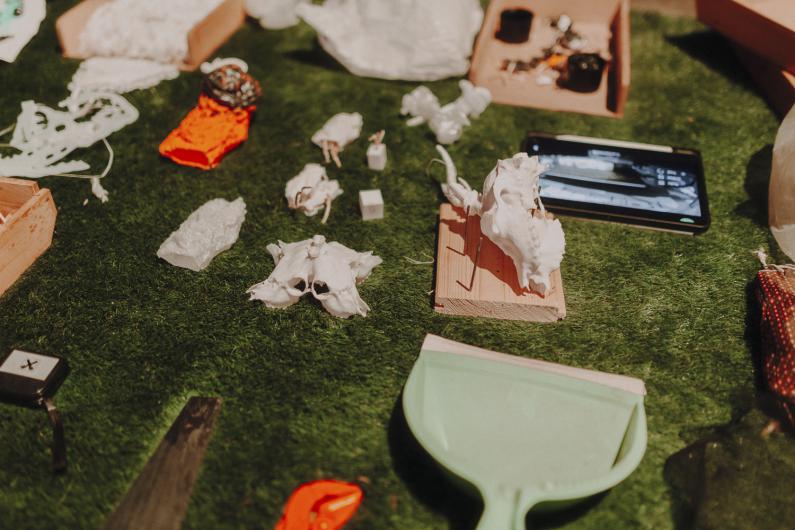 Close-up of green artificial grass with various white objects such as cast bones, small sculptures, wooden blocks, a smartphone and tools scattered across the surface.