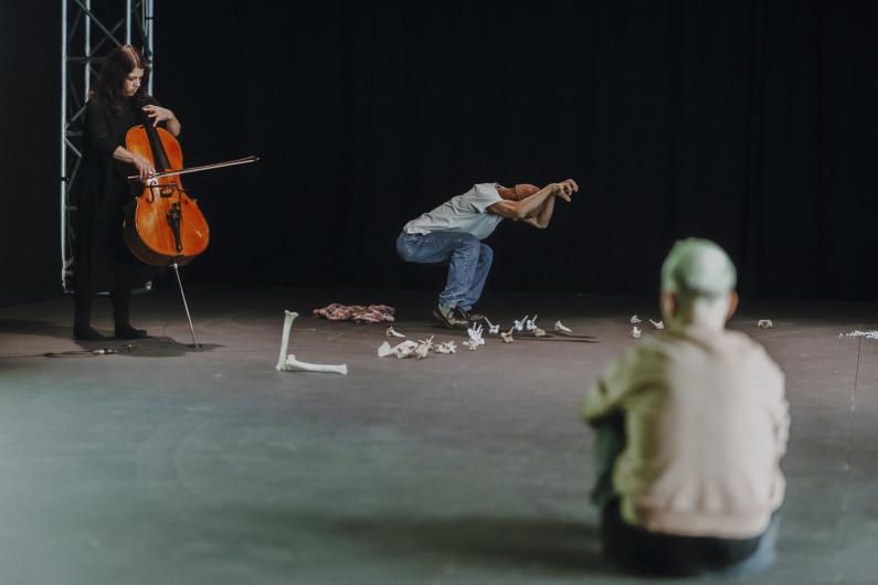 Stage with a black backdrop: on the left a person plays the cello, in the middle a person in jeans crouches in a sculptural pose, in the foreground another person sits on the floor watching, with bone-like objects scattered between them.