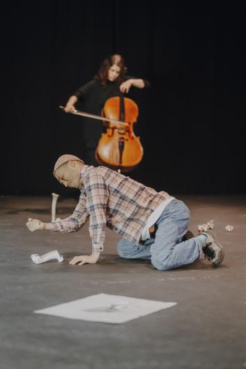 A person in a checked shirt kneels on the stage floor and bends toward a bone object, while a cellist plays in the background against a black curtain.