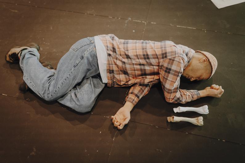 A person in jeans and a checked shirt lies curled on their side on the stage floor with hands clenched, three bone objects placed in a row in front.