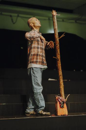 A person in a checked shirt and jeans stands on a tiered platform, looking up at an upright tree trunk with cut branches that is mounted on a painted wooden block.