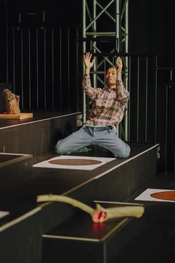 A person in a checked shirt kneels on a tiered platform, raising both arms upward with a focused expression, while a small plinth with a piece of wood sits beside them.