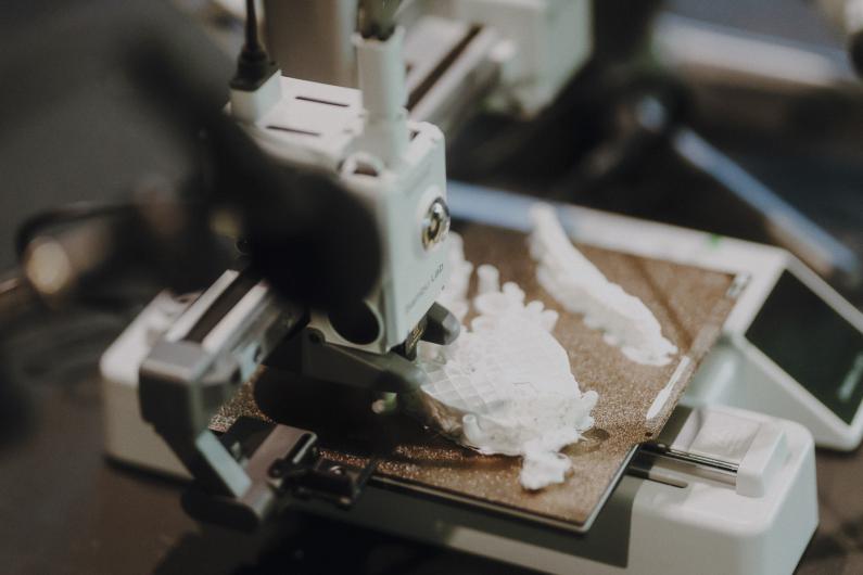 Close-up of a 3D printer producing white, organic-looking forms on its print bed, with cables and machine parts filling the frame.