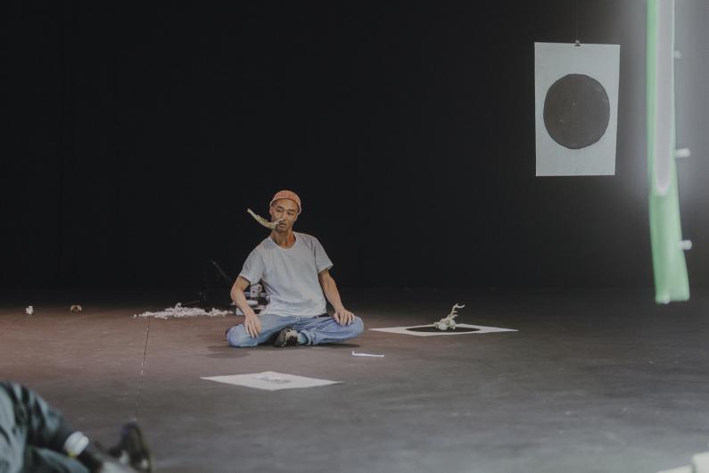 A person sits cross-legged on stage with a thin piece of wood or bone held between their lips, while a sheet with a black circle and parts of the installation hang behind them.