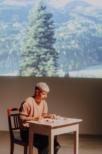 A person with short light hair sits at a wooden table, carefully working on small objects, while a blurred projection of a mountain forest with a fir tree appears in the background.