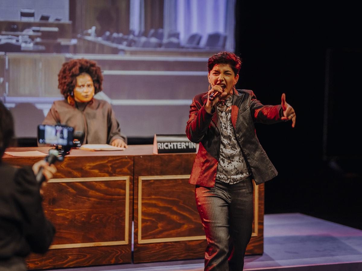 In the foreground, a person performs energetically with a microphone. Behind them, another person sits at a desk labeled “Judge.”