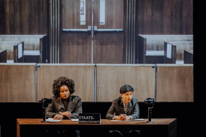 Two people sit at a desk labeled “Prosecution.” Both wear formal clothing, cameras record the scene, and a wooden courtroom backdrop is visible behind them.