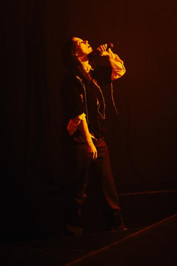 A performer stands alone on stage in warm orange light. The person wears a dark top layered over a light long-sleeved shirt, wide dark trousers, and light sneakers. Holding a microphone, they tilt their head back with their mouth open, as if speaking or singing. The background is dark, and the lighting creates strong contrasts.