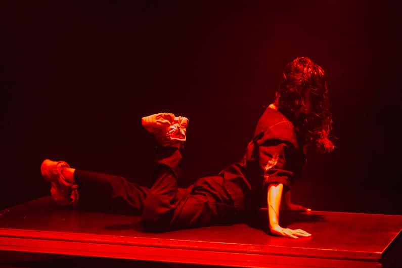 A performer lies on their stomach on a low rectangular stage platform. The lighting is intense red. They support themselves with one hand; one leg is bent, showing a light-colored sneaker. Loose dark clothing with light fabric details drapes softly. The scene feels powerful and physically expressive.