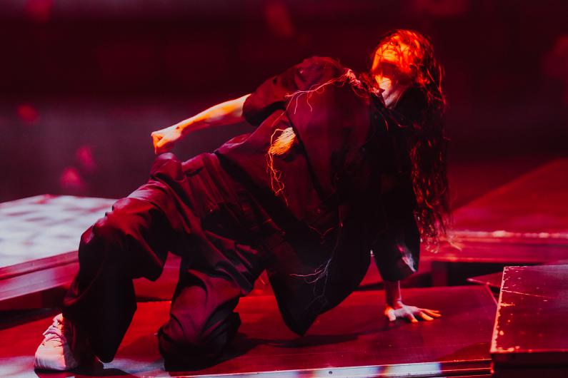 Eine Performer*in kniet seitlich auf einer Plattform, der Oberkörper ist zurückgelehnt, eine Faust geballt. Langes, dunkles Haar fällt nach hinten. Rotes Bühnenlicht modelliert Gesicht und Kleidung. Mehrere versetzte Podeste bilden eine geometrische Spielfläche im Hintergrund.