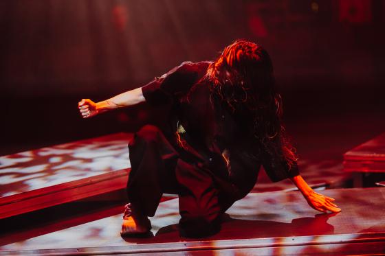 A performer sits low on a platform, supporting themselves with one arm behind them while the other hand forms a fist. Their face is partly covered by hair. Red light dominates the scene, while bright spots cast patterns onto the stage floor.