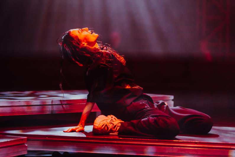 A performer leans back against a platform, head tilted far back. Warm red light illuminates their face and upper body. Their legs are bent, and a light sneaker rests visibly on the stage. The pose appears open and intense.