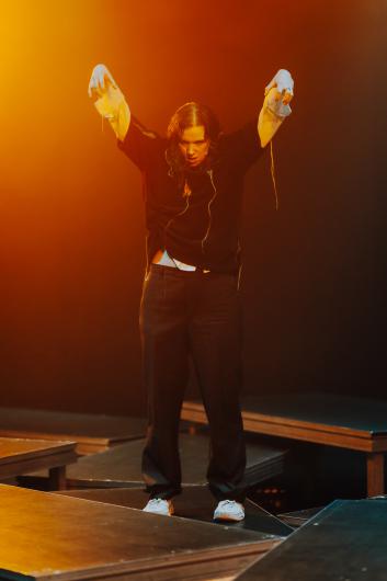 A performer stands on a platform with both arms raised, hands loosely curved. The lighting shifts between warm orange and cooler tones. Several low platforms are arranged at angles, creating a layered stage landscape.