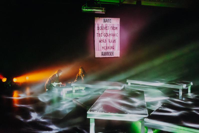 Zwei Performer*innen befinden sich auf einer Bühne mit mehreren Podesten. Nebel und farbige Lichtstrahlen durchziehen den Raum. Im Hintergrund hängt ein Banner mit englischem Text: „RAPE DERIVES FROM THE GERMANIC WORD ‘RAUB’ MEANING ROBBERY.“ Die Szene verbindet Performance, Lichtinstallation und Text.