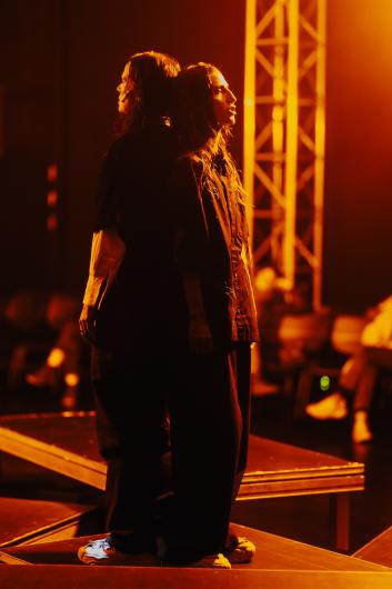Two performers stand back to back on a platform. Warm orange light illuminates their profiles. In the blurred background, audience seating and stage structures are visible. The scene conveys both tension and connection.