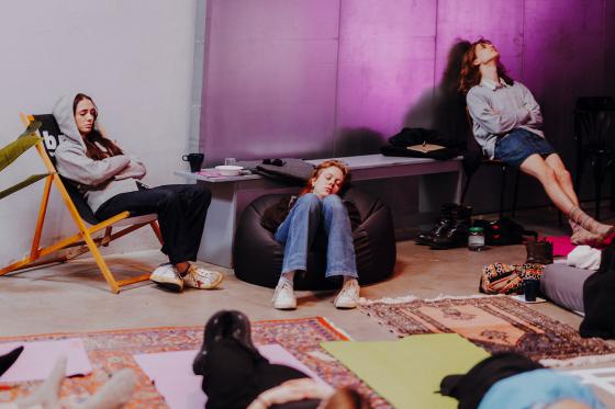 Several people are sitting or lying relaxed in a room with rugs and mats. One person sits in a deck chair wearing a hoodie, another in a beanbag, and a third leans back on a chair with their head tilted up. The room is bathed in warm purple light, creating a calm, meditative atmosphere.