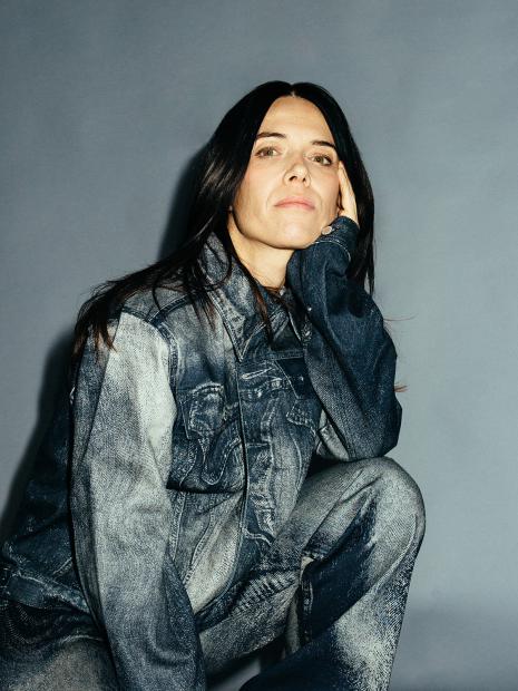 A person with long dark hair looks calmly at the camera, resting their head on one hand. They are wearing a denim outfit with a washed texture. The background is solid gray.