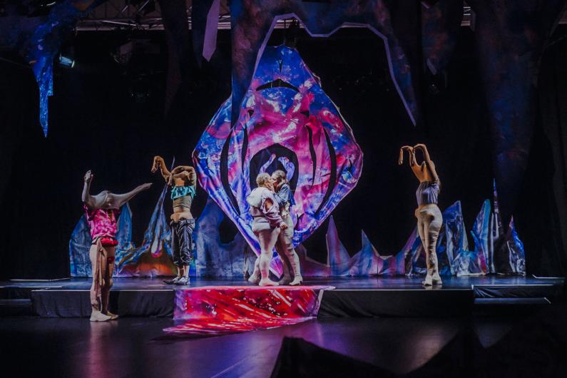 Mehrere Performer*innen stehen und bewegen sich auf einer Bühne vor einer großen, abstrakten Bühneninstallation in Blau-, Pink- und Lilatönen. In der Mitte stehen zwei Personen einander sehr nahe, während andere seitlich mit erhobenen Armen oder verhüllten Köpfen agieren.