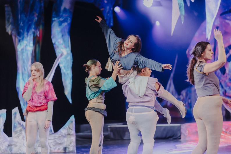 Eine Person wird von mehreren Performer*innen angehoben und getragen. Die getragene Person streckt einen Arm nach oben, der Körper ist waagrecht ausgerichtet. Die umstehenden Personen stützen den Körper mit Händen und Armen. Im Hintergrund hängen abstrakte Bühnenelemente.