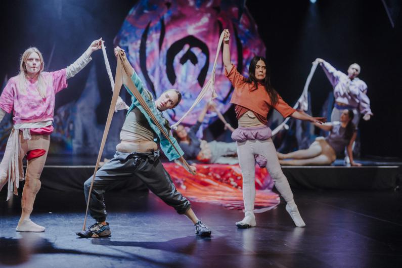 Mehrere Performer*innen stehen in einer Reihe auf der Bühne und halten lange, elastische Stoffstreifen in den Händen. Die Körper sind unterschiedlich geneigt, die Arme gespannt. Im Hintergrund ist eine große, organisch wirkende Bühnenform in kräftigen Farben zu sehen.