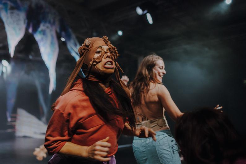 Eine Person trägt eine Stoffmaske, die das Gesicht teilweise verhüllt, und bewegt sich mit ausdrucksstarker Mimik nach vorne. Eine weitere Person tanzt dahinter. Das Bühnenlicht ist gedämpft, mit Nebel und kühlen Farbtönen.