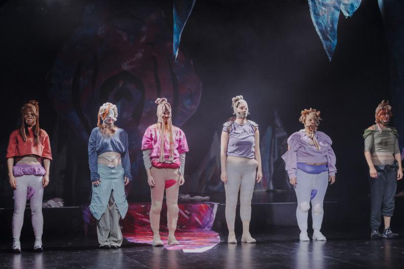 Eine Gruppe von Performer*innen steht nebeneinander in einer Reihe, alle mit Stoffen oder Masken am Kopf oder Körper. Die Körperhaltung ist aufrecht und ruhig. Das Licht fällt frontal auf die Gruppe, der Hintergrund bleibt dunkel.