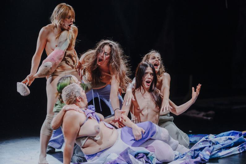 Eine Gruppe von Performer*innen liegt und kniet eng beieinander auf einer Stoffbahn am Bühnenboden. Mehrere Personen strecken die Arme nach oben, Münder sind geöffnet, die Körper wirken angespannt und expressiv. Das Licht ist dunkel mit starken Kontrasten und betont Haut, Stoffe und Bewegung.