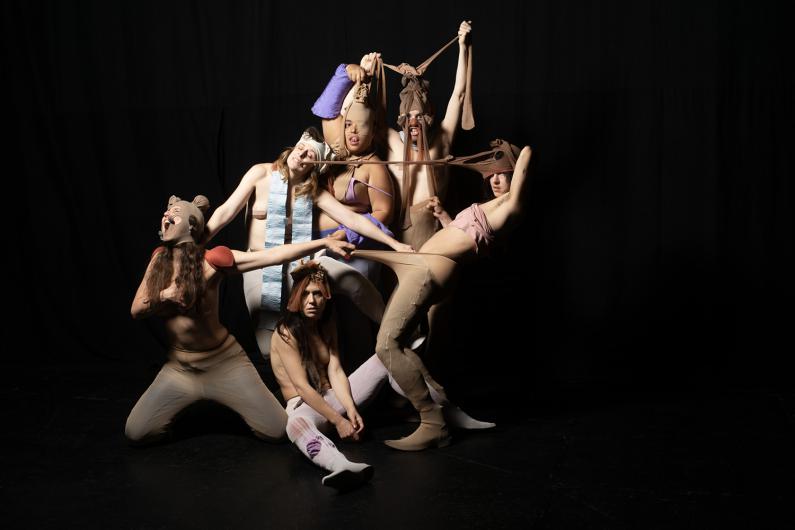 The group of performers poses in a sculptural composition. Tights are stretched over heads and bodies, creating a humorous and surreal scene.