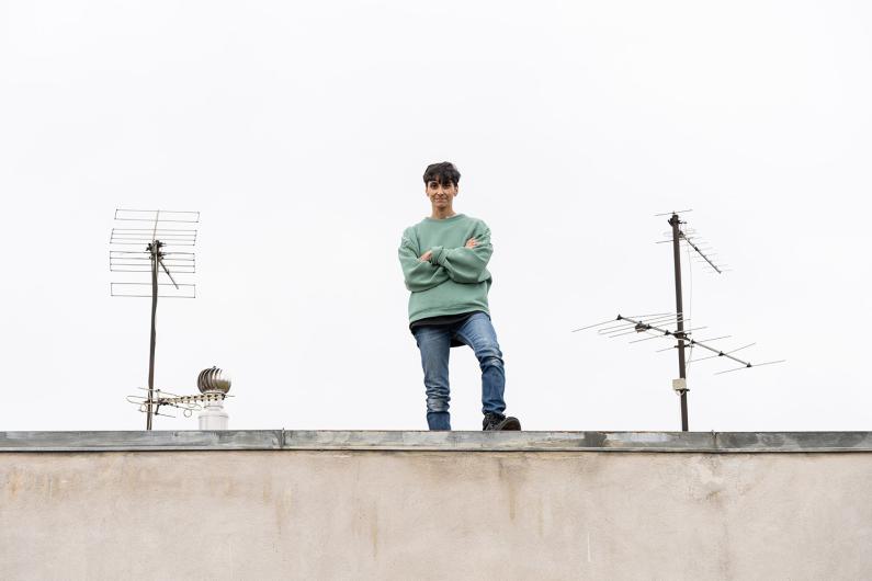 Eine Person in Jeans und grünem Pullover steht mit verschränkten Armen auf einem Dach zwischen zwei Fernsehantennen, vor grauem Himmel.